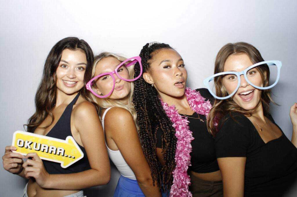 Four friends posing joyfully in a photobooth with oversized colorful glasses and playful props, showcasing their bond and the fun atmosphere of the event.