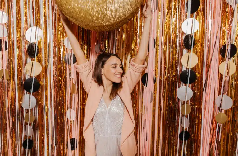 Woman in a sparkling dress and pink blazer posing with a glittering photo booth backdrop of gold, pink, and black streamers.