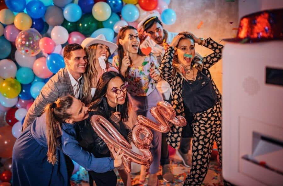 Group of friends enjoying a colorful photo booth rental with props and balloons.