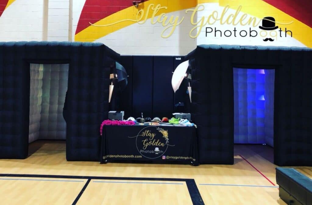 Stay Golden inflatable photo booth setup in a gym with props table between two black booths.