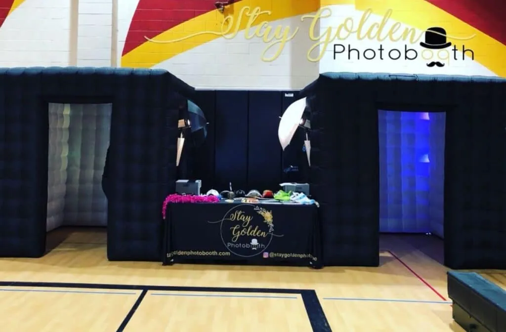 Stay Golden inflatable photo booth setup in a gym with props table between two black booths.