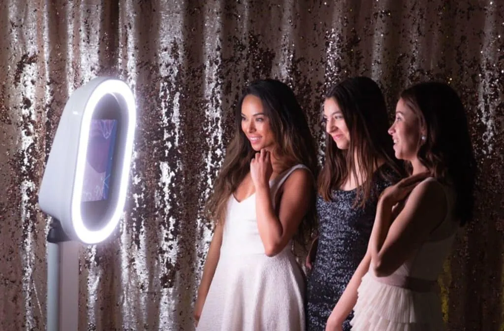 Three friends smiling and posing at a classic photo booth with a LED light against a sequined backdrop.