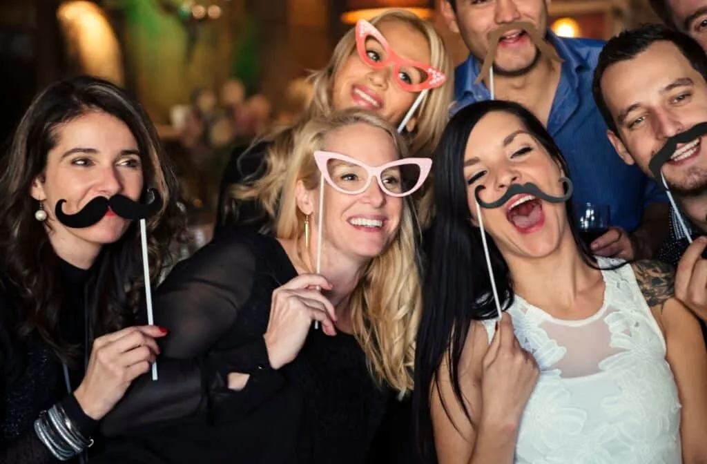 Group of friends laughing and posing with mustache and glasses props in a photo booth.