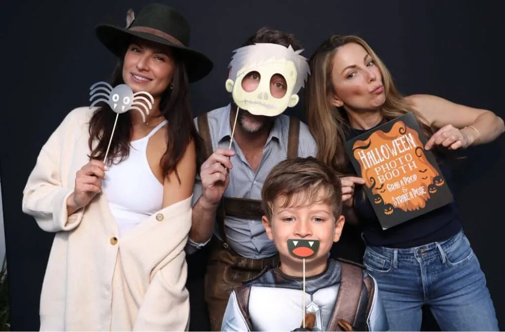 Family posing with Halloween photo booth props, including masks, signs, and spider decor.