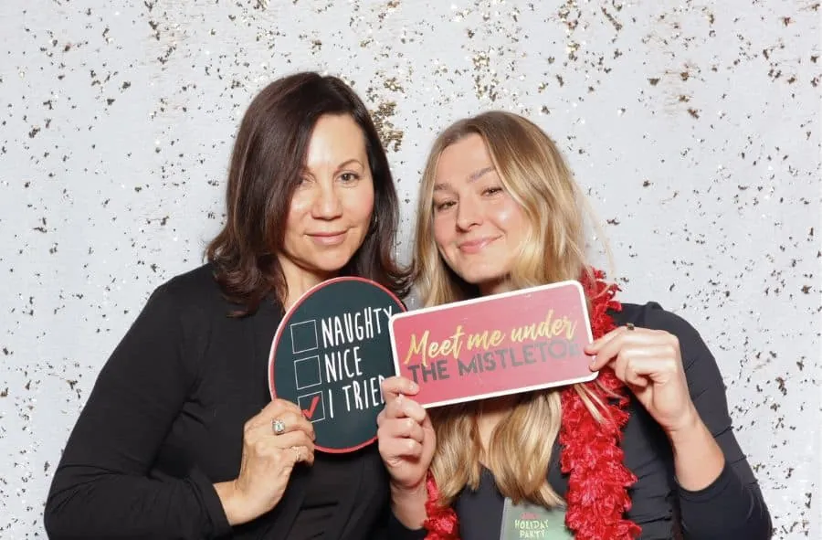 Two women posing with Christmas-themed photo booth props, including "Naughty" and "Mistletoe" signs.