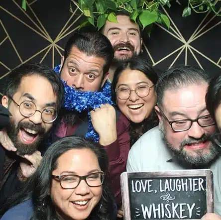 A group of friends posing in a photo booth with props, laughter, and a "Love, Laughter, and Whiskey" sign.