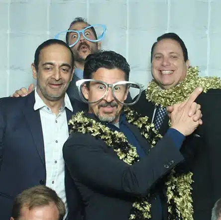 A group of men posing playfully in a photo booth, wearing oversized glasses and gold tinsel scarves.