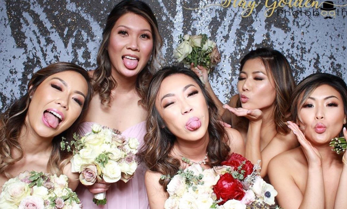 Bridesmaids pose playfully with bouquets at a wedding photo booth with a sparkling backdrop.