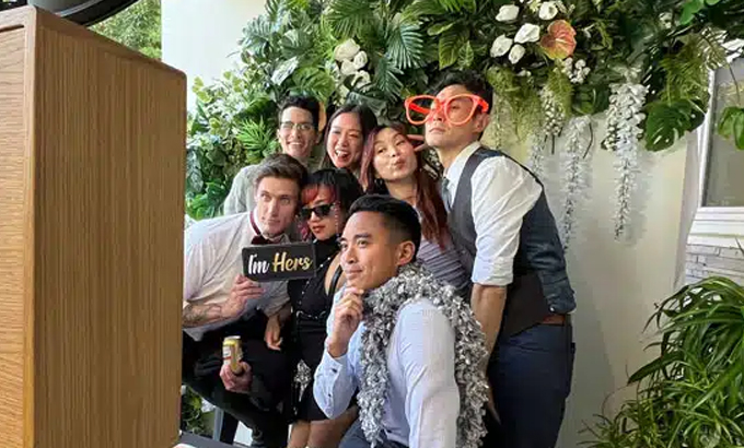 A group of friends and relatives pose playfully with props at a wedding photo booth with a floral backdrop.