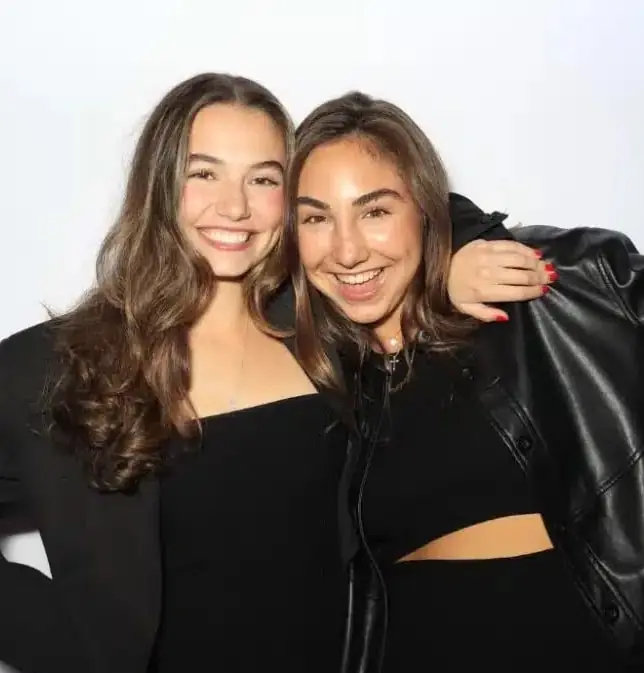 Two smiling friends in black outfits pose together at a party photo booth rental with a simple white backdrop.