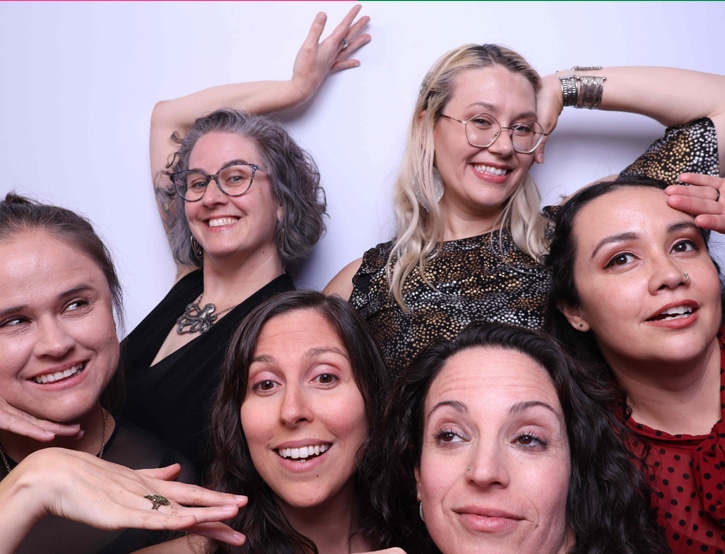 Six women strike playful poses at a corporate photo booth with a simple white backdrop.