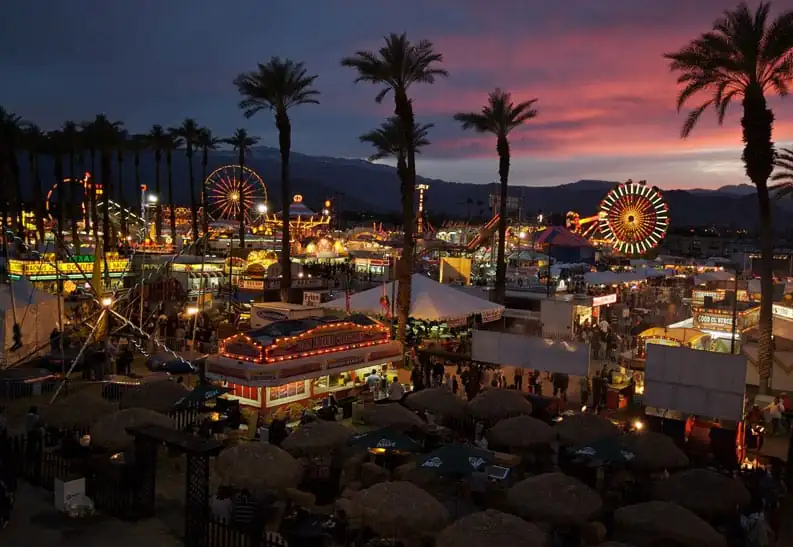 A lively fair in Riverside County at sunset, featuring Ferris wheels, food stands, and palm trees.