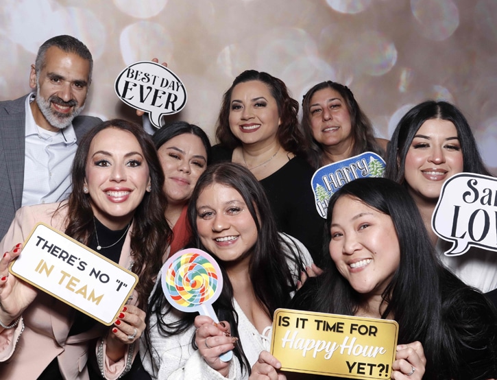 A group of coworkers poses with playful props at a corporate photo booth with a bokeh backdrop.