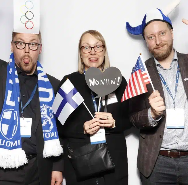 Three people pose with Finnish and U.S. flags, props, and playful expressions at a corporate event.