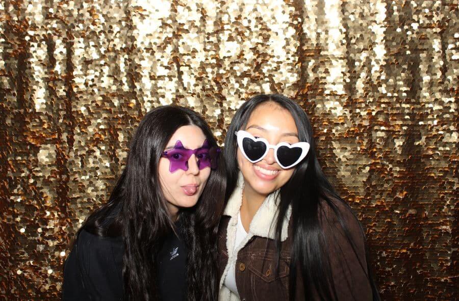 Two women wearing star and heart sunglasses smiling in front of a gold photo booth backdrop.