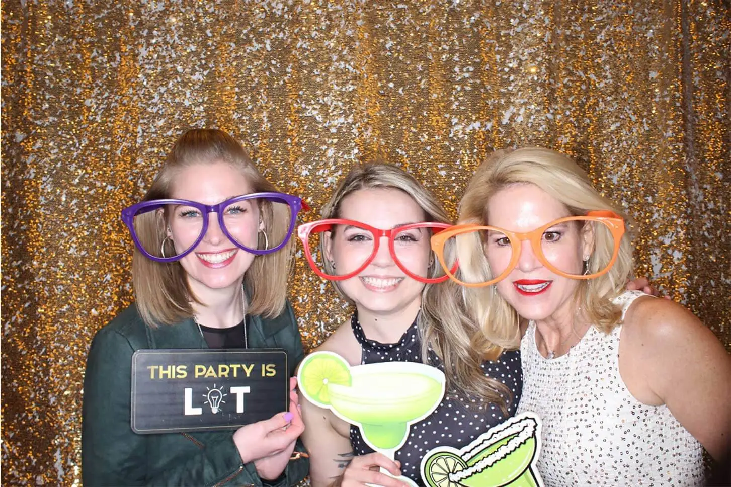 Three ladies pose with oversized glasses in front of a gold sequin backdrop at a mini photo booth.