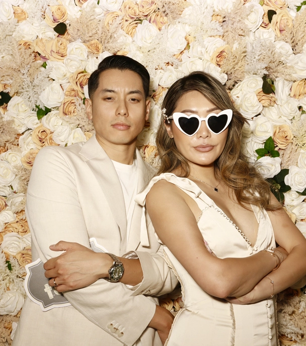 Stylish newlyweds in matching outfits pose confidently at a wedding photo booth with a floral backdrop.