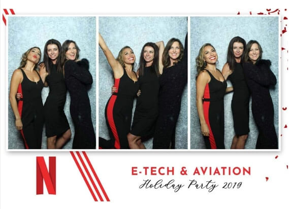 Group posing in a mirror photo booth at a festive corporate holiday party with a silver backdrop.