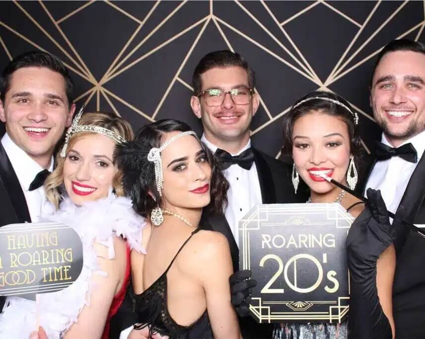 Group dressed in Roaring 20s theme posing in classic photo booth with vintage backdrop and props.