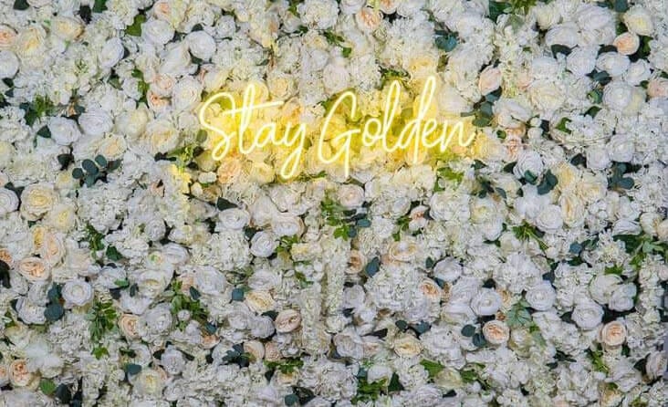 Glam Garden photo booth flower wall with white roses and a glowing "Stay Golden" neon sign.