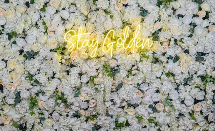 Glam Garden photo booth flower wall with white roses and a glowing "Stay Golden" neon sign.