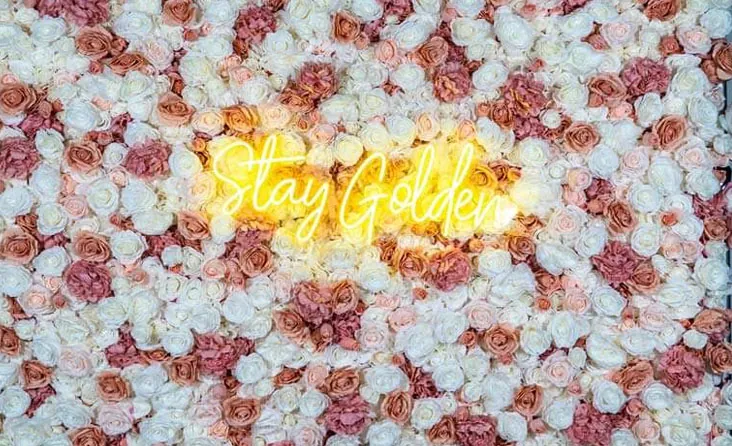 Blushing photo booth flower wall with pink, white, and peach roses and "Stay Golden" neon sign.