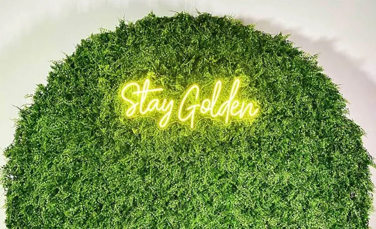 Hedge-style photo booth backdrop with dense greenery and a glowing "Stay Golden" neon sign.