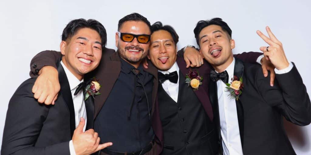 Four men in suits posing playfully at The Rico Wedding's photo booth rental in Newport Beach, flashing smiles.