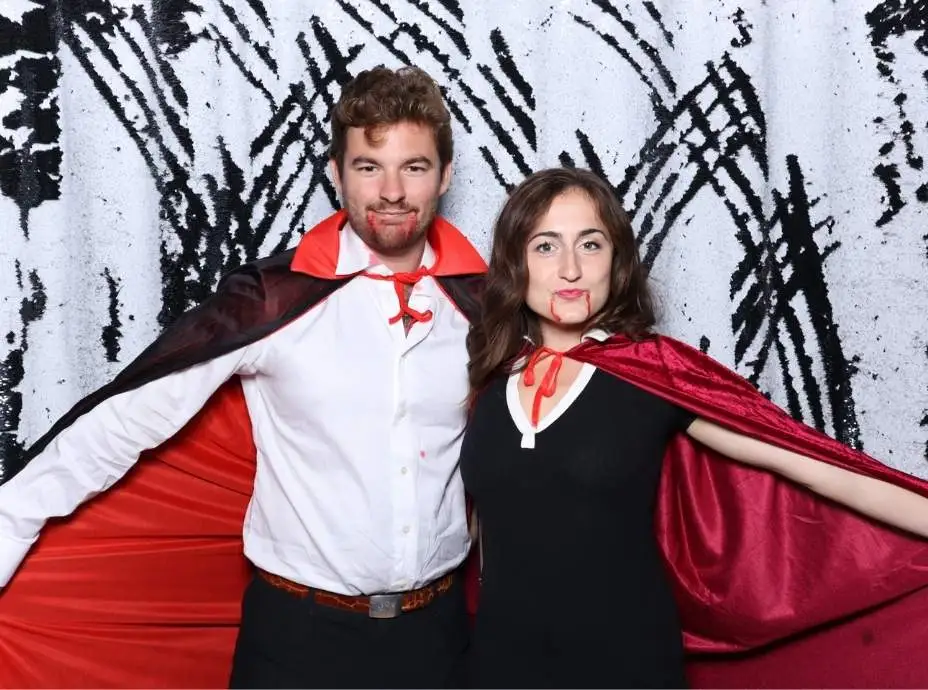 Couple dressed as vampires wearing red capes and fangs posing dramatically in a Halloween photo booth.