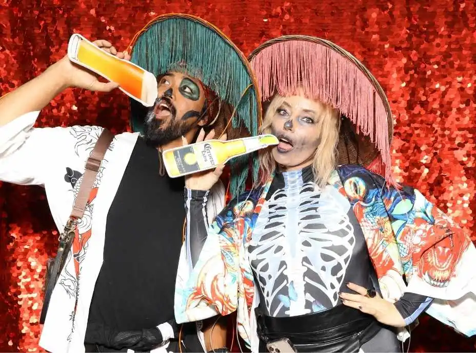 Couple in vibrant Day of the Dead costumes posing with sombreros and drink props in a photo booth.