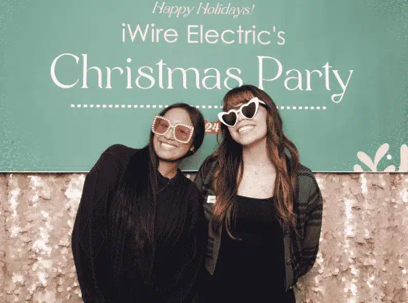 Two ladies smiling in front of a branded backdrop wearing fun holiday glasses at iWire Electric's christmas party
