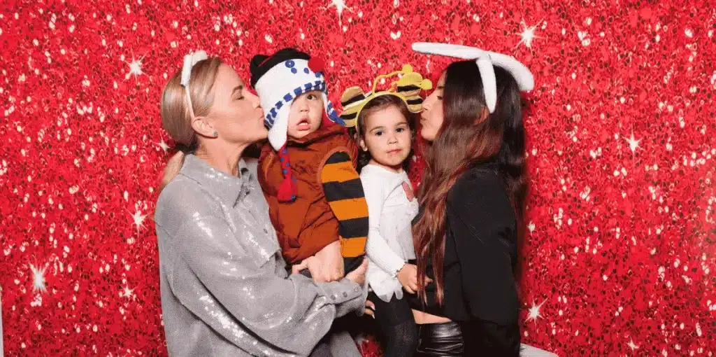 Two moms holding toddlers with fun Christmas props in front of a red sparkling holiday backdrop