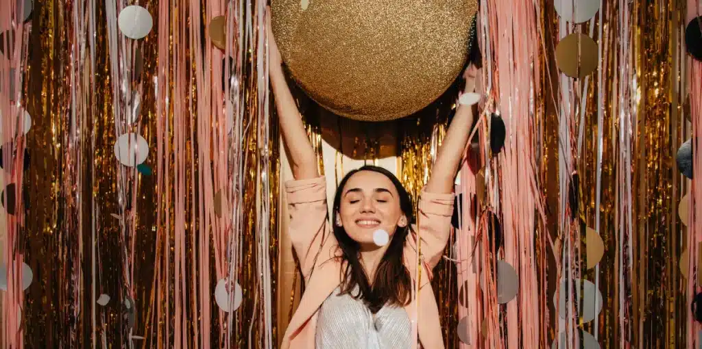 A lady holding up a gold ball in front of a gold and pink sequin backdrop for new year's eve photo booth