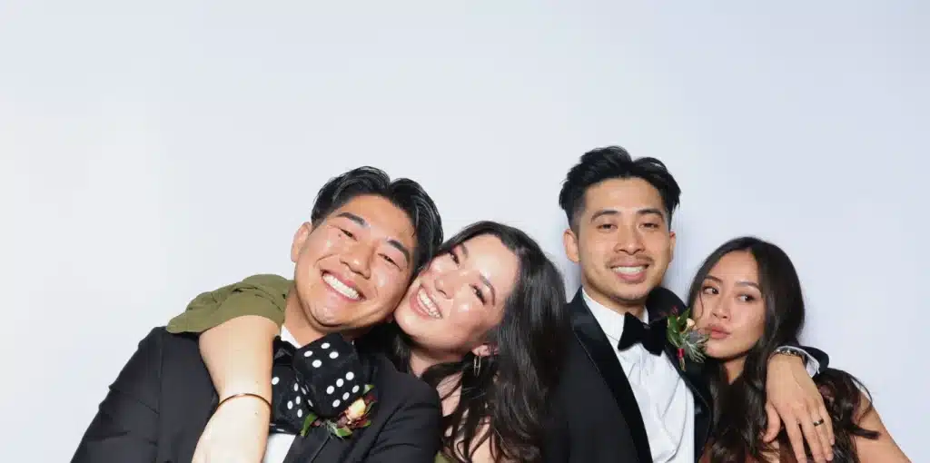 Four smiling people dressed in formal attire pose closely together against a white background.