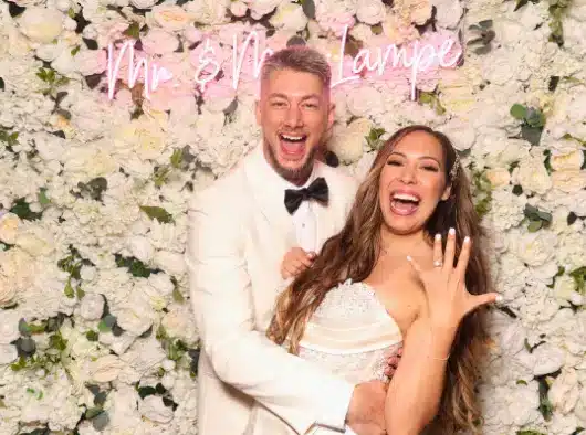 A couple poses in front of a floral wall with a neon sign that reads "Mr. & Mrs. Lampe"; one person is wearing a white suit and the other is wearing a wedding dress and showing off a ring.