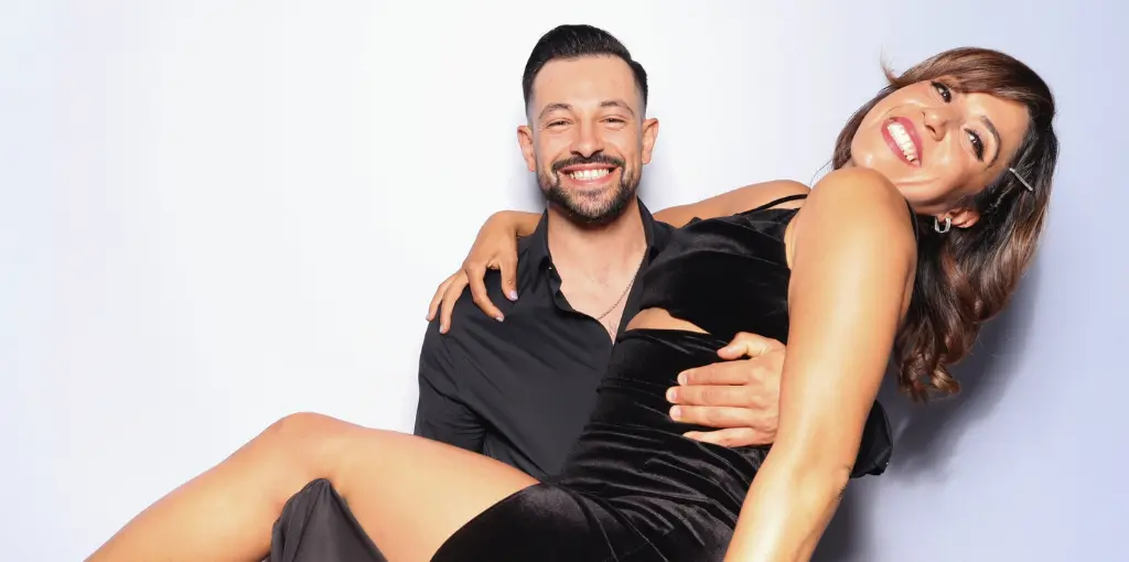 A man in a black shirt holds a woman in a black dress, both smiling against a white background.