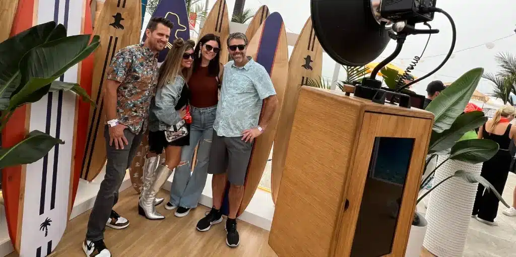 A group of four people pose for a photo in front of a backdrop of surfboards and tropical plants, with a wooden photo booth to the right.