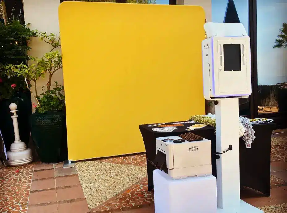 A classic photo booth with a screen and blue LED lights stands on a white pedestal next to a table with props, in front of a bright yellow backdrop. A printer sits on a white cube to the left of the photo booth.