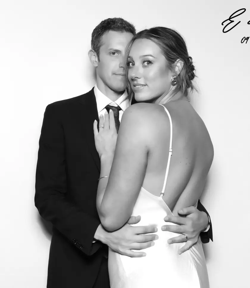 A couple poses for a glam photo booth. The person on the left wears a suit and tie, while the person on the right wears a white dress. Their arms are around each other.
