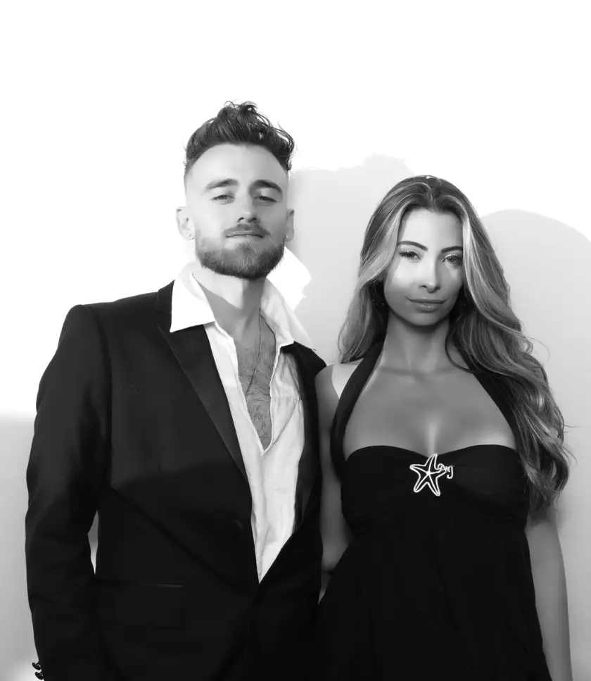 A man in a suit and an unbuttoned shirt stands next to a woman in a black dress with a star embellishment on the bodice.