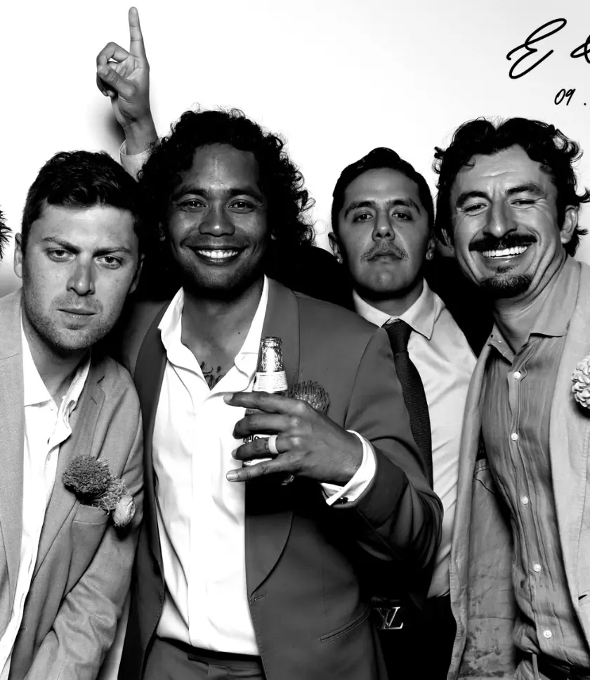 Four men in formal wear pose for a wedding photo booth, with one person holding a bottle and another pointing upwards.
