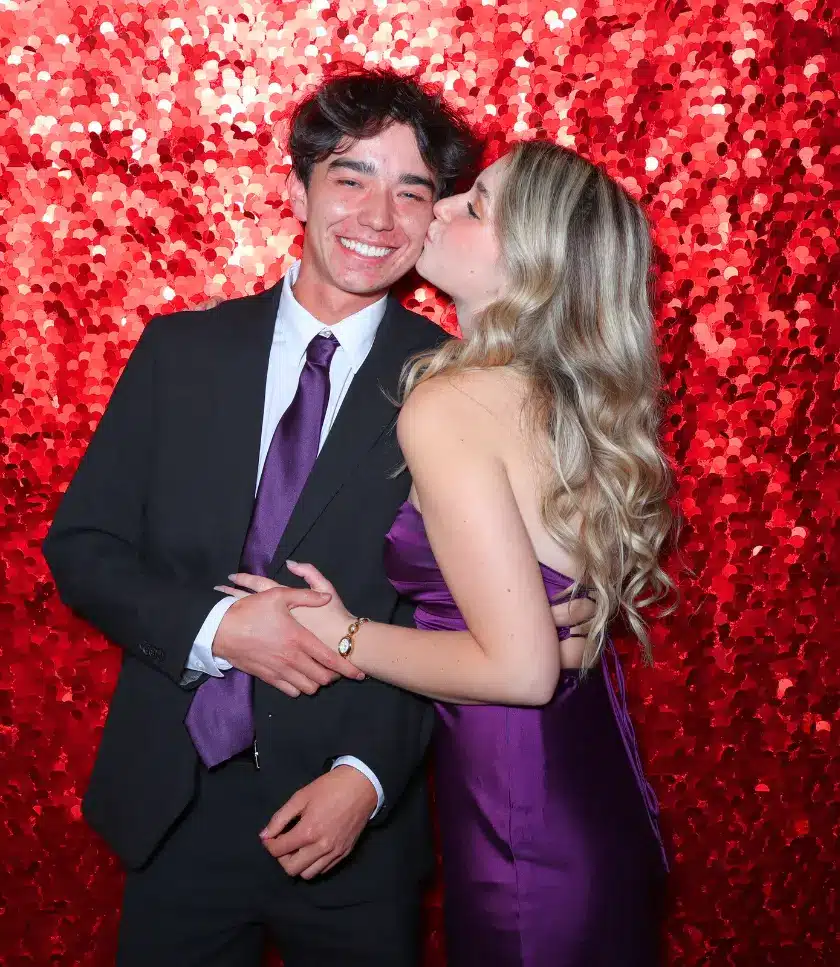 A person in a suit and tie is kissed on the cheek by a person in a purple dress, in front of a red sequined backdrop.