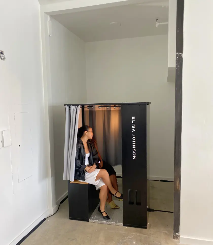 Two people sit inside an enclosed photo booth with "ELISA JOHNSON" printed vertically on the side.