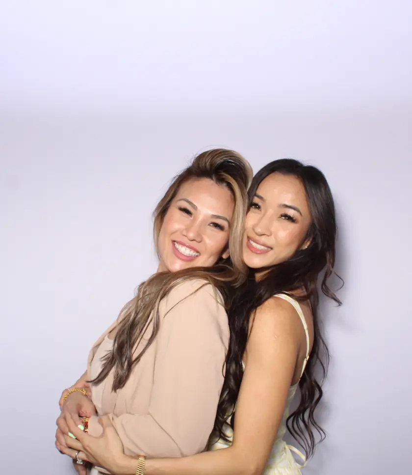 Two pretty ladies embrace against a white backdrop, both smiling warmly at the photo booth