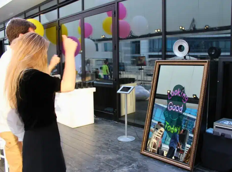 A mirror photo booth with the words "Looking Good Today" written on it in neon-like lettering, with a ring light attached to the top. Two people are visible in the reflection, and balloons are hanging in the background.