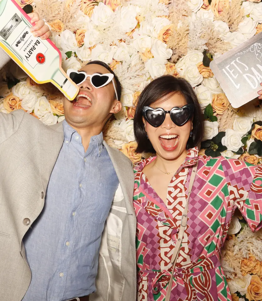 Two people wearing heart-shaped sunglasses pose for a photo in front of a floral backdrop. One person holds up a cutout of a Bacardi bottle, while the other holds a sign that says "Let's Dance".