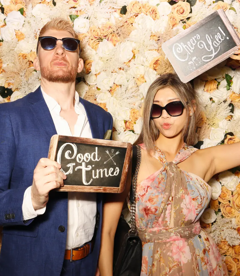 A man in a blue suit and sunglasses holds a chalkboard sign that reads "Good Times". A woman in a floral dress and sunglasses stands next to him, holding a sign that reads "Cheers". They are posing in front of a floral backdrop.