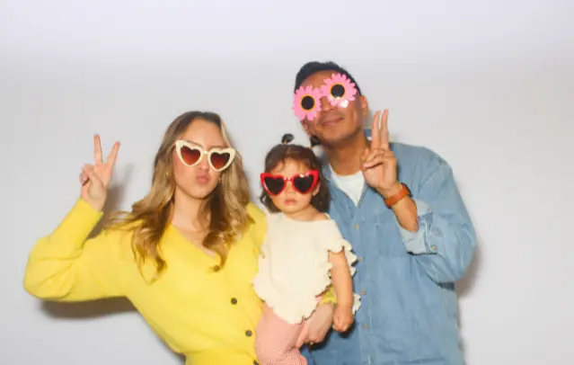 A family of three, wearing heart-shaped sunglasses and flower-shaped glasses, pose for a photo against a white background, making peace signs with their hands.