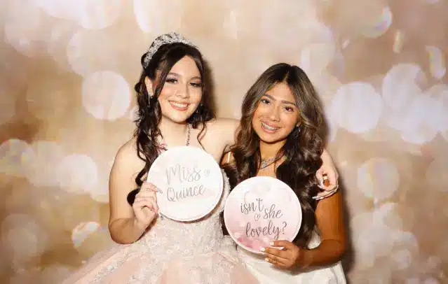 A person in a tiara and a pink dress holds a "Miss Quince" sign, while another person in a white dress holds a "Isn't she lovely?" sign, with a bokeh background.