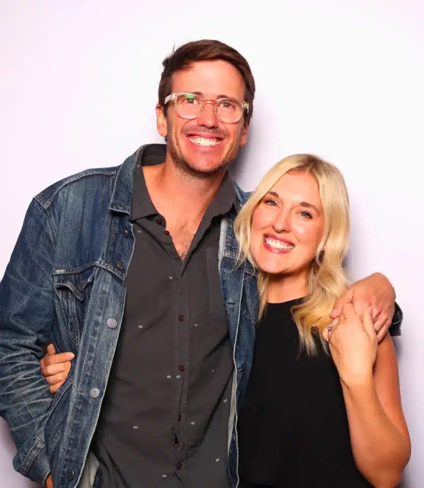 A smiling parent-to-be are wearing glasses and a denim jacket stands with an arm around another smiling individual in a black top against a white background.